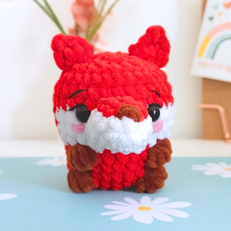 May include: A handmade, red crocheted fox with white and brown accents. The plush toy has black eyes and pink cheeks, and is sitting on a light blue surface with a daisy pattern. The fox has a cute, cartoonish appearance.