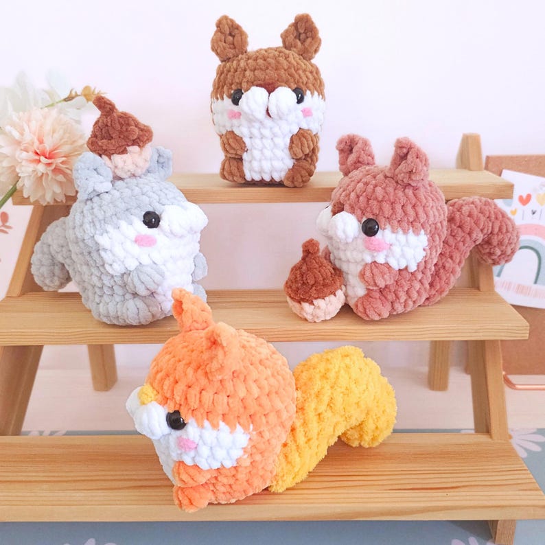 Op de afbeelding: Vier handgemaakte gehaakte eekhoornknuffels in bruin, grijs, oranje en roze. Elke eekhoorn heeft een witte buik, een kleine eikel en zwarte ogen. De knuffels worden tentoongesteld op een houten plank.