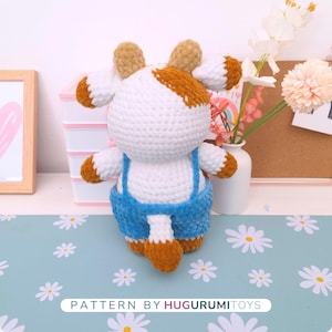 May include: A handmade crochet cow plushie with white, brown, and blue accents. The cow has a white body, brown ears and patches, and is wearing blue overalls. The text "PATTERN BY HUGURUMITOYS" is at the bottom.