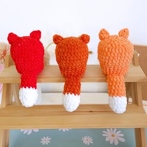May include: Three handmade crochet fox toys in vibrant colors. The foxes have orange, red, and light orange bodies with white-tipped tails. Each fox has small, pointed ears and a fluffy texture. They are displayed on a wooden shelf.
