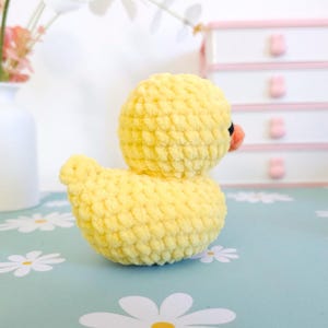 Puede incluir: Un patito amarillo tejido a mano con ganchillo, con cabeza y cuerpo redondos. El patito tiene un peque&ntilde;o pico naranja y ojos negros. La lana suave y texturizada le da al pato una apariencia tierna. El pato est&aacute; sobre una superficie azul claro con patrones de margaritas blancas.