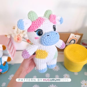 May include: A handmade, plush cow toy with a white body and colorful accents. The cow features pink, blue, green, and yellow details, including spots and ear tips. The text "PATTERN BY HUGURUMITOYS" is visible at the bottom of the image.