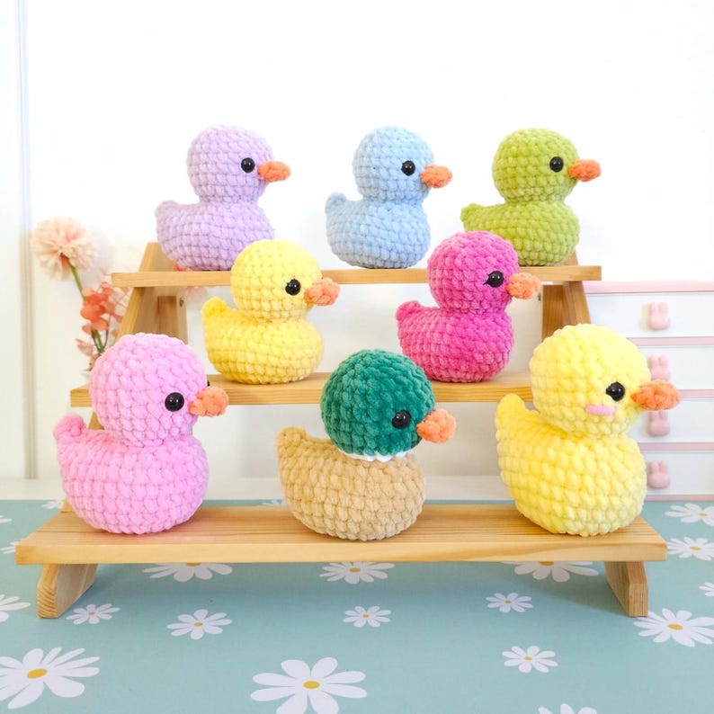 Puede incluir: Una colecci&oacute;n de patitos de ganchillo hechos a mano en varios colores, incluyendo rosa, amarillo, azul, verde y morado. Cada patito tiene un peque&ntilde;o pico naranja y ojos negros. Los patitos est&aacute;n expuestos en una estanter&iacute;a de madera.