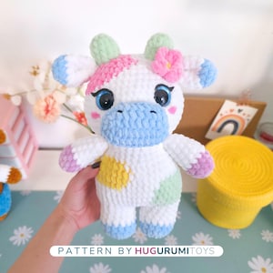 May include: A handmade white crochet cow plushie with blue, pink, yellow, and green accents. The cow has large blue eyes, a pink flower, and the text "PATTERN BY HUGURUMITOYS".