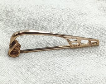 May include: A gold-colored brooch pin with a safety pin design. The pin features a coiled spring at one end and a decorative, geometric pattern near the clasp. The brooch is displayed on a textured, off-white surface.