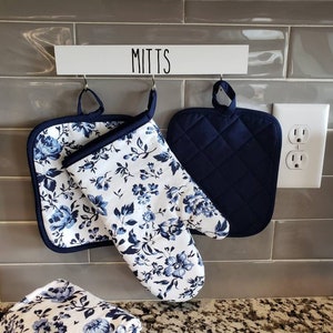 May include: A set of kitchen mitts and a pot holder hanging on a wall. The mitt and pot holder feature a white background with blue floral patterns. A solid navy blue pot holder and a sign that reads "MITTS" are also visible.