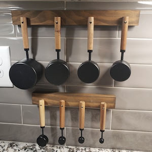 May include: A set of six black measuring cups with wooden handles hanging on a wooden rack. The cups are arranged in a row, with the handles facing the viewer. The rack is mounted on a white tiled wall.