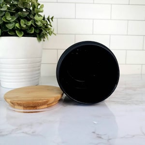Pode incluir: Um pote de cer&acirc;mica preto com tampa de bambu. O pote est&aacute; vazio e a tampa est&aacute; ao lado dele sobre uma superf&iacute;cie de m&aacute;rmore branco.