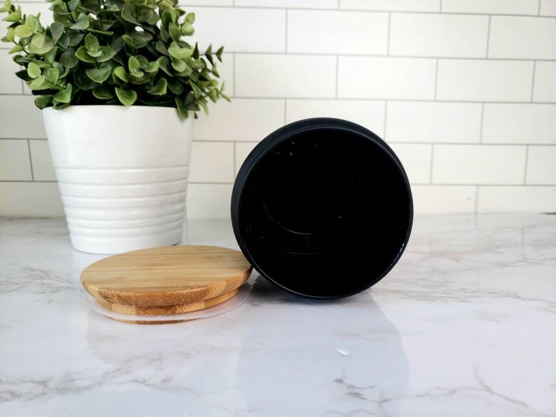 Pode incluir: Um pote de cer&acirc;mica preto com tampa de bambu. O pote est&aacute; vazio e a tampa est&aacute; ao lado dele sobre uma superf&iacute;cie de m&aacute;rmore branco.
