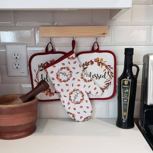 May include: Two red and white oven mitts with the words "blessed" and "grateful" hanging on a wooden rack. A wooden mortar and pestle sits on the counter next to a bottle of olive oil.