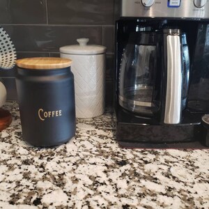 Pode incluir: Um pote de caf&eacute; de cer&acirc;mica preto com uma tampa de madeira e a palavra "COFFEE" em letras douradas. O pote est&aacute; sentado em uma bancada ao lado de um pote de cer&acirc;mica branco e uma cafeteira preta.