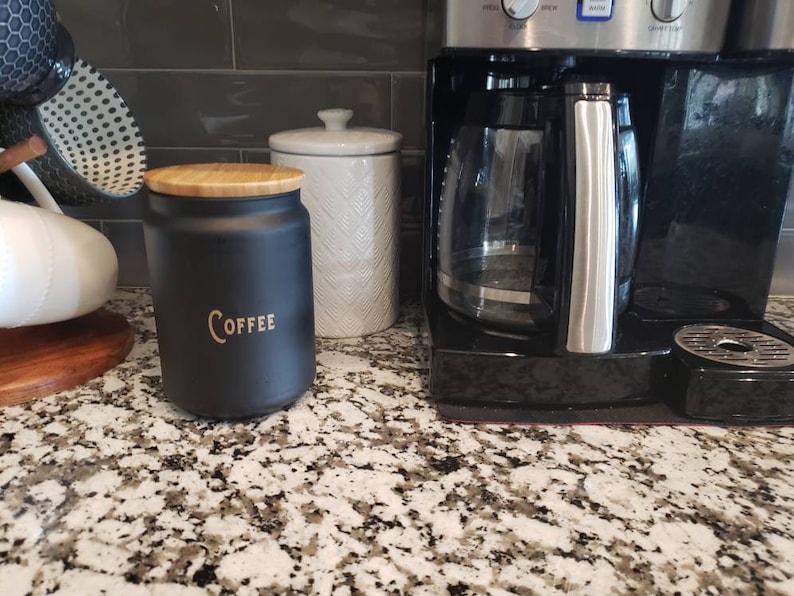 Pode incluir: Um pote de caf&eacute; de cer&acirc;mica preto com uma tampa de madeira e a palavra "COFFEE" em letras douradas. O pote est&aacute; sentado em uma bancada ao lado de um pote de cer&acirc;mica branco e uma cafeteira preta.