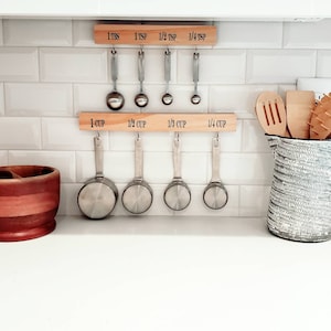 May include: A wooden rack with metal measuring cups and spoons hanging from it. The rack has labels for 1 tbs, 1 tsp, 1/2 tsp, 1/4 tsp, 1 cup, 1/2 cup, 1/3 cup, and 1/4 cup. The rack is mounted on a white tiled wall.