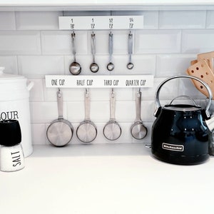 May include: A white kitchen counter with a black electric kettle, a white ceramic canister labeled "Flour", two spice shakers labeled "Salt" and "Pepper", and a set of measuring cups and spoons hanging on a white rack. The rack has labels for "One Cup", "Half Cup", "Third Cup", and "Quarter Cup".