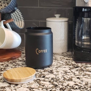 Pode incluir: Um pote de caf&eacute; de cer&acirc;mica preto com uma inscri&ccedil;&atilde;o dourada que diz "COFFEE" e uma tampa de bambu. O pote est&aacute; em cima de uma bancada de granito.