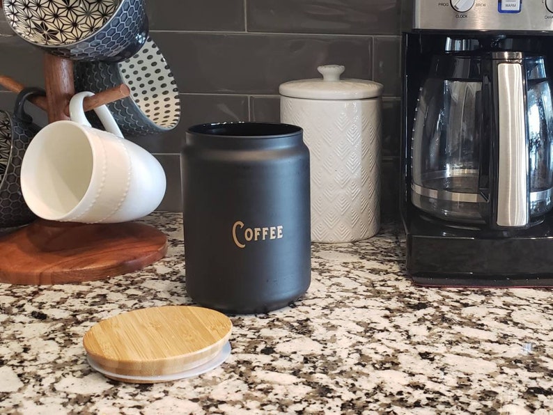 Pode incluir: Um pote de caf&eacute; de cer&acirc;mica preto com uma inscri&ccedil;&atilde;o dourada que diz "COFFEE" e uma tampa de bambu. O pote est&aacute; em cima de uma bancada de granito.