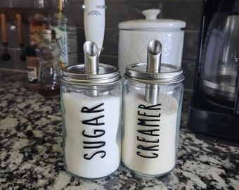 May include: Two glass jars with metal lids and spouts, labeled "Sugar" and "Creamer". The jars are filled with white sugar and cream, respectively. The jars are sitting on a speckled countertop.