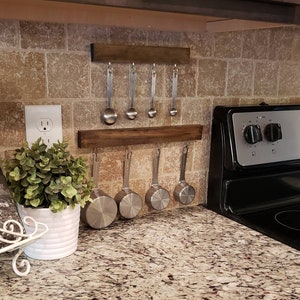 May include: A wooden kitchen utensil rack with metal hooks holding measuring spoons and cups. The rack is mounted on a tiled wall above a granite countertop.