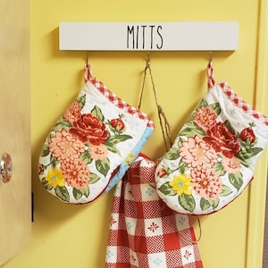 May include: A white wooden rack with the word "MITTS" written on it. Two floral oven mitts and a red and white checkered tea towel are hanging from hooks on the rack.