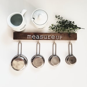 May include: A wooden rack with the text "measure up" painted in white, hanging on a white wall. Four silver measuring cups hang from hooks on the rack.