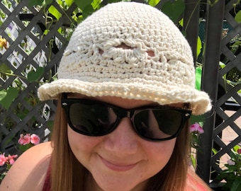 May include: A woman wearing a white crocheted sun hat with a wide brim and black sunglasses. She is smiling and looking at the camera.