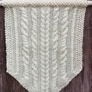 May include: Cream-colored, knitted wall hanging with a cable knit design. The banner is rectangular with a pointed bottom and is suspended from a wooden dowel. The textured pattern features vertical cable and ribbed sections.