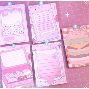 Op de afbeelding: Set van zes roze en witte notitieblokken met verschillende illustraties. De illustraties omvatten een retro arcade-spel, een smartphone, een donutwinkel, een bloedzak en een hamburger. De tekst op de notitieblokken is "Catcher", "Yum!" en "Tactoki Illustrations".