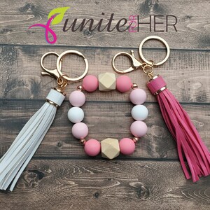 May include: A pink and white beaded bracelet with wooden beads and gold keychains with tassels. The bracelet is attached to the keychains. The keychains have white and pink tassels.