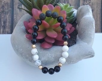 May include: A black and white beaded bracelet with gold accents. The bracelet features black lava beads, white marble beads, and gold spacer beads.