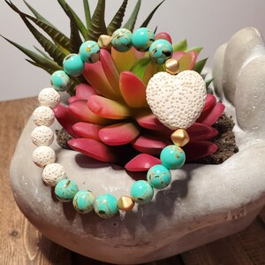 May include: A turquoise and white beaded bracelet with a white heart-shaped lava stone charm. The bracelet is made with small round beads and gold spacers.
