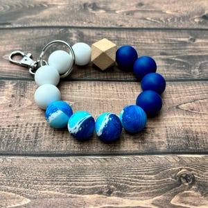 May include: A blue and white silicone beaded keychain with a wooden bead and a silver key ring. The beads are in a gradient pattern, starting with white, then blue with white swirls, and finally solid blue.