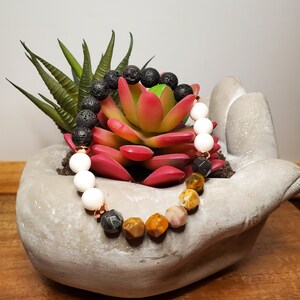 May include: A bracelet made with black lava rock, white beads, and tiger's eye, rose quartz, and golden yellow beads. The bracelet is displayed on a concrete hand-shaped planter with a succulent plant.