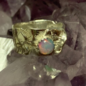 Könnte beinhalten: Ein silberner Ring mit einem strukturierten Band und einem runden Opal-Edelstein in der Mitte. Der Opal ist schillernd in Rosa-, Grün- und Blautönen.