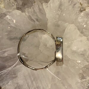 May include: A silver ring with a smooth, oval-shaped stone set in the center. The ring is displayed on a bed of white crystals.