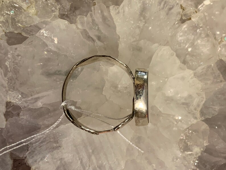 May include: A silver ring with a smooth, oval-shaped stone set in the center. The ring is displayed on a bed of white crystals.