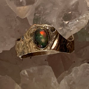 Puede incluir: Un anillo de plata con una banda texturizada y una gran piedra preciosa de ópalo ovalada. El ópalo muestra un juego de colores con tonos verdes, rojos y negros. El anillo tiene un aspecto rústico y artesanal.