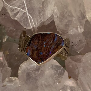 May include: A silver ring with a dark blue and brown opal gemstone set in a silver setting. The ring is displayed on a bed of white crystals.