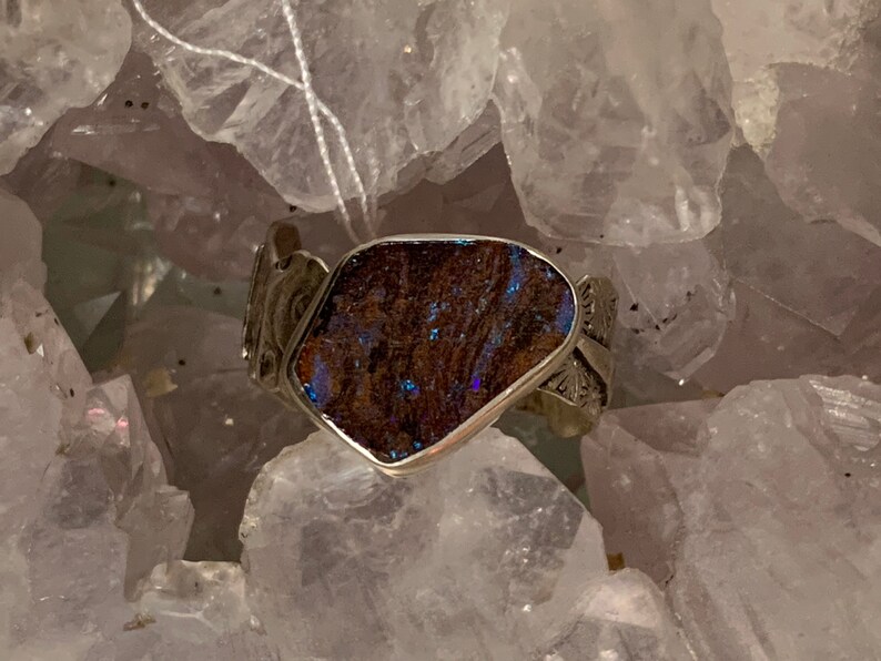 May include: A silver ring with a dark blue and brown opal gemstone set in a silver setting. The ring is displayed on a bed of white crystals.