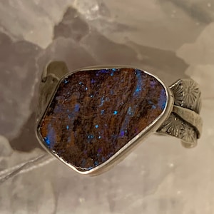 Puede incluir: Un anillo de plata con una gran piedra de ópalo de forma irregular engastada en el centro. El ópalo tiene una base marrón con destellos azules y verdes. La banda del anillo está decorada con un diseño floral.
