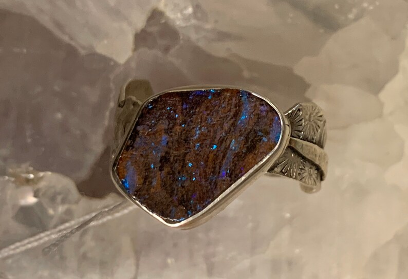 May include: A silver ring with a large, irregular-shaped opal stone set in the center. The opal has a brown base with blue and green flashes of color. The band of the ring is decorated with a floral design.