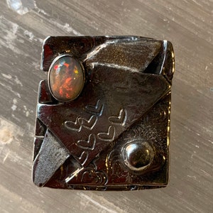 Puede incluir: Un anillo de plata con una piedra de ópalo negro y un diseño de corazón. El anillo está hecho de metal y tiene una superficie texturizada.
