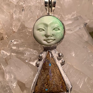 May include: A silver pendant with a carved green moon face and a triangular piece of brown opal. The pendant is set against a background of white quartz crystals.