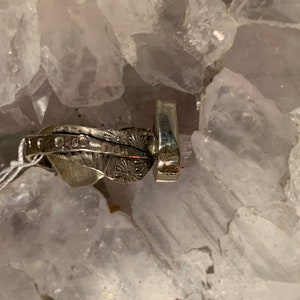 May include: A silver ring with a textured design and a small, round stone set in the center. The ring is on a bed of white crystals.