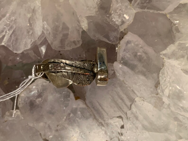May include: A silver ring with a textured design and a small, round stone set in the center. The ring is on a bed of white crystals.
