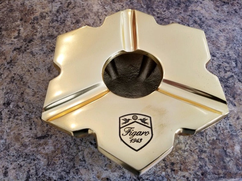 Puede incluir: Un cenicero dorado con forma de estrella y un logotipo que dice "Figaro 1943".