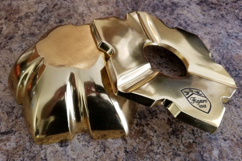 Puede incluir: Un cenicero de metal dorado con un dise&ntilde;o decorativo. El cenicero tiene una abertura redonda en el centro y un borde elevado. El cenicero est&aacute; marcado con el texto "Figaro 1943".