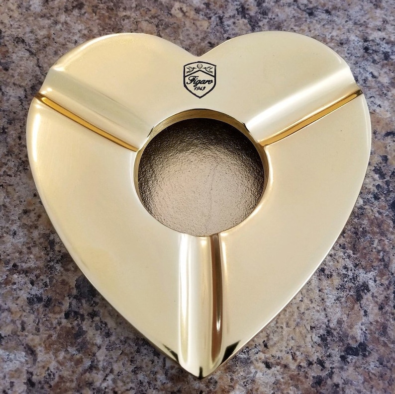 May include: A gold heart-shaped ashtray with three compartments and a round center. The ashtray has the text "Figaro 1943" in a shield-shaped logo.