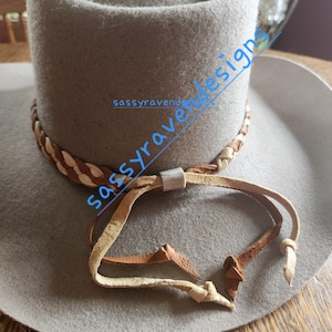 May include: A light gray cowboy hat with a braided leather band and a leather chin strap. The hat has a wide brim and a tall crown. The chin strap is made of light brown leather and is attached to the hat with a metal slider. The text "sassyravendesigns" is written on the hat.