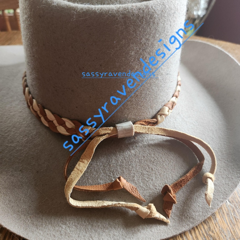 May include: A light gray cowboy hat with a braided leather band and a leather chin strap. The hat has a wide brim and a tall crown. The chin strap is made of light brown leather and is attached to the hat with a metal slider. The text "sassyravendesigns" is written on the hat.