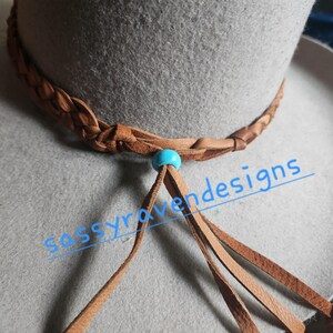 May include: Close-up of a gray hat with a braided brown leather band. A turquoise bead and brown leather tassels hang from the band. The text "sassyravenesigns" is visible in the image.